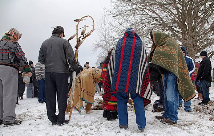 Tribal members gathered outdoors in winter, several wrapped in traditional blankets, during a ceremony